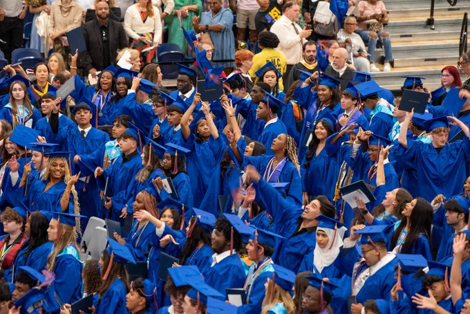 Large group of graduates wearing blue robes and caps, posing for a photograph outdoors.
