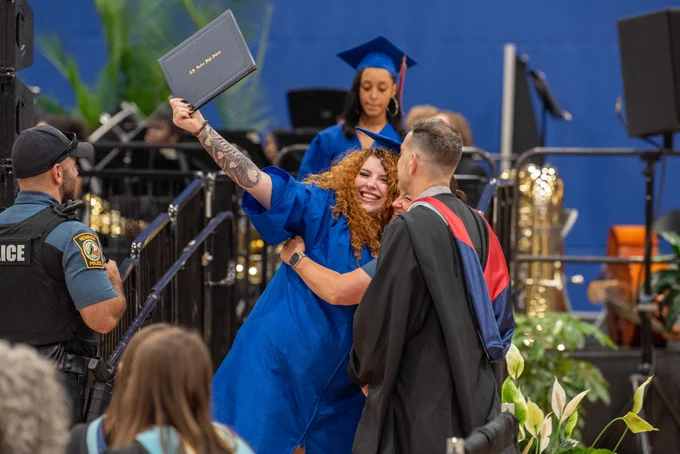 A graduate in a blue cap and gown standing on a stage, holding a diploma, with a police officer nearby and spectators in the background.