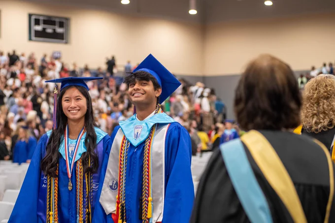 A graduation ceremony with two smiling students in blue graduation gowns and caps standing together.