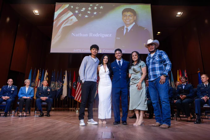 A group of people stands on a stage during a ceremony. One person is wearing a uniform, receiving an honor or award from another individual. A large screen in the background displays the name "Nathan Rodriguez" and mentions achievements in criminal justice. Several flags are visible in the background.