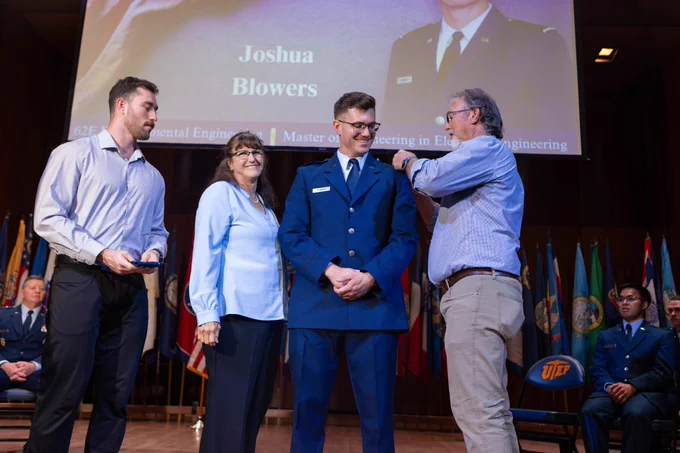 A group of people stands on a stage during a ceremony. One person is wearing a uniform, receiving an honor or award from another individual. A large screen in the background displays the name "Joshua Blowers" and mentions achievements in engineering. Several flags are visible in the background.