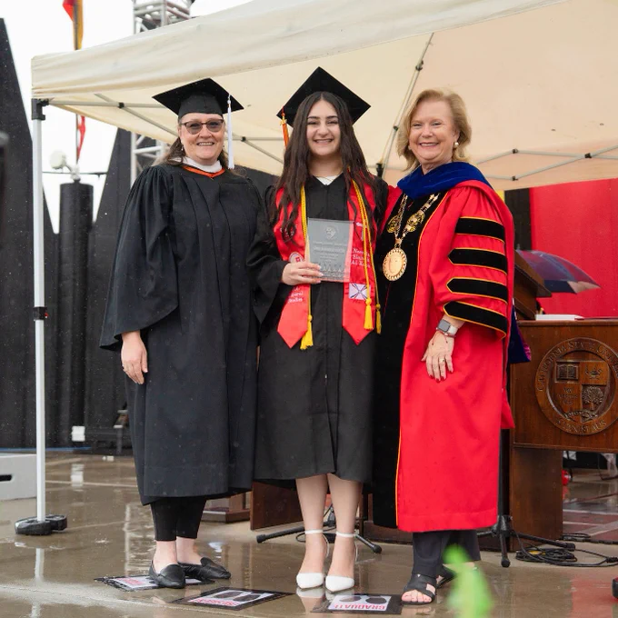 Three women in graduation attire stand smiling under a tent. The graduate in the center holds a plaque, flanked by two faculty members in black and red robes.