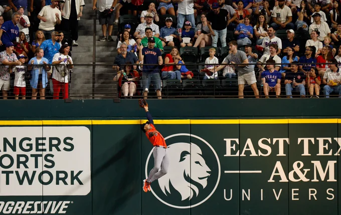 Cam Smith leaps at the wall to rob a home run during Thursday’s game. He wears an orange Astros uniform and blue cap while fans in the stands look on.