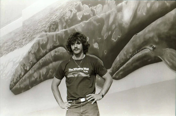 Black and white photo of artist Robert Wyland standing in front of mural of two whales.