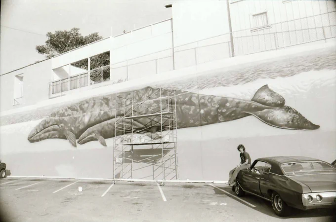 Black and white photo of mural on wall of two whales. Scaffolding structure placed next to the wall. Robert Wyland is sitting on the hood of a car parked in the lot near the end of the whale mural.
