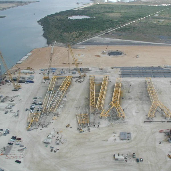 Aerial view of Kiewit's Offshore Services Yard showing several large yellow structures, cranes and vehicles on a sandy expanse near a waterway.
