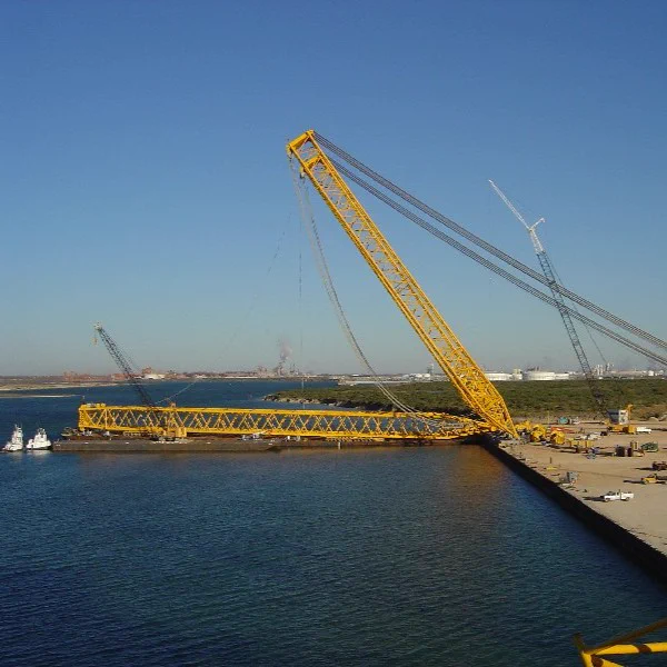 A large yellow crane on a dock protrudes over water, with an expansive industrial area in the background. Several smaller cranes and equipment are visible on land. The sky is clear and blue.