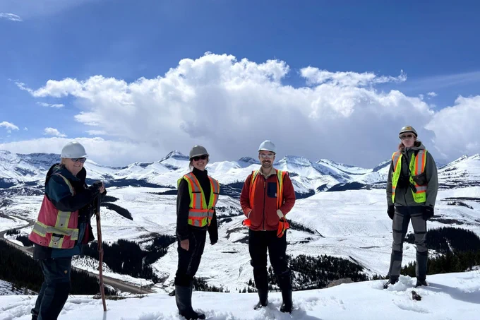 Four biologists in PPE stand on a snowy hilltop with mountains in the background
