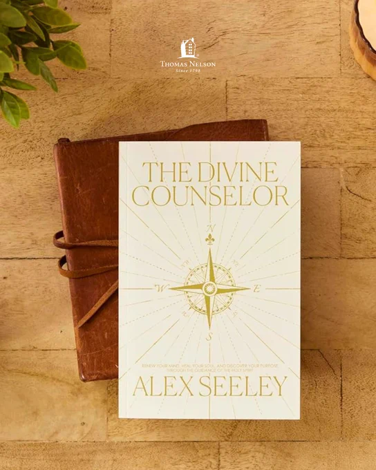 A book titled "The Divine Counselor" by Alex Seeley is placed on top of a closed, brown leather journal on a wooden surface. The book cover features an ornate compass design. In the upper left corner, there's a small plant. The publisher, Thomas Nelson, is noted above the book.
