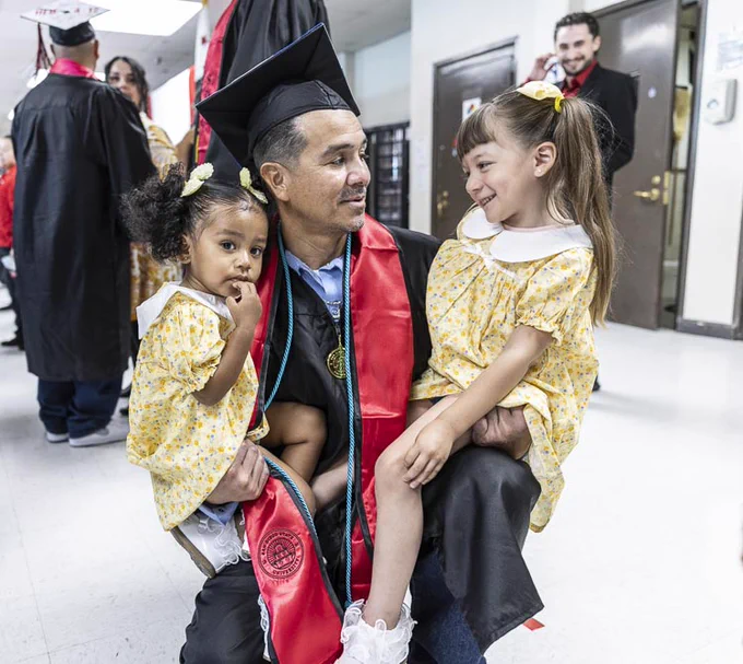 A person in a graduation gown and cap is kneeling, holding two young children in matching yellow dresses. They are in a hallway where others in graduation attire are visible. The scene appears celebratory and joyful.