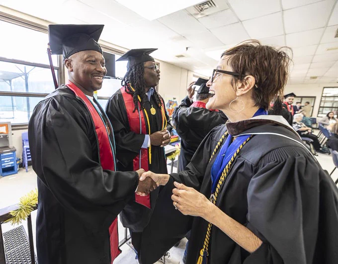 Graduates in caps and gowns are shaking hands with a faculty member inside a classroom. The setting is lively and celebratory, with people smiling and engaging warmly.