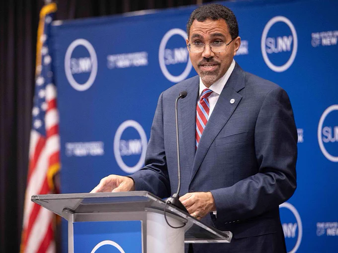 Person standing at a podium, speaking in front of a blue backdrop with the SUNY logo. An American flag is positioned to the side.