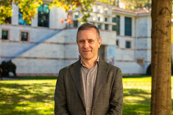 Environmental portrait of Benjamin Reiss in a dress shirt and jacket standing on the Emory Quad, with the Carlos Museum and campus greenery blurred in the background
