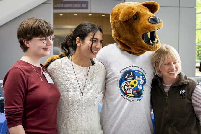 A group of four people: three in casual attire and one in the Nittany Lion costume. The mascot wears a T-shirt. Another person wears a vest with the WWF logo. They are standing closely together, smiling in a bright indoor setting.