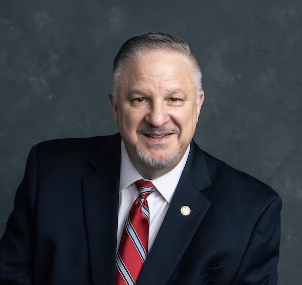 Richard Tyler, CEO of Richard Tyler International, Forbes Business Council member, award‑winning filmmaker, Broadway producer, and founder of Richard Tyler Films and Excellence Edge Films, photographed in formal business attire with a dark suit, white shirt, and red, white, and blue striped tie against a textured dark gray background. A circular lapel pin featuring his trademarked Commitment to Excellence philosophy is prominently displayed.
---
