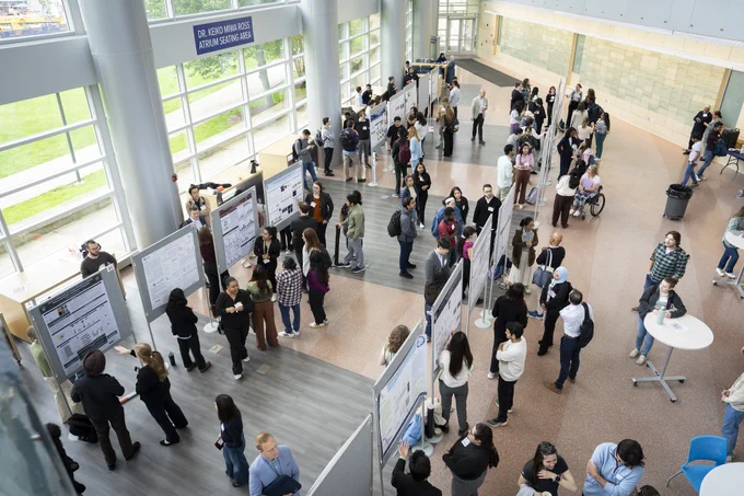 A busy science poster session. Attendees are engaged in discussions around various research posters displayed on boards. The atrium's large windows let in ample natural light. People are gathered in groups, some standing at tables, others examining the posters closely.