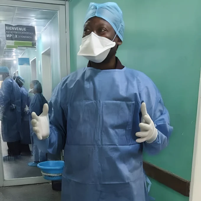 A person in medical scrubs, mask, gloves, and a hair covering is standing in a hospital corridor. They appear to be speaking or explaining something. A sign on the wall reads "BIENVENUE" in French.