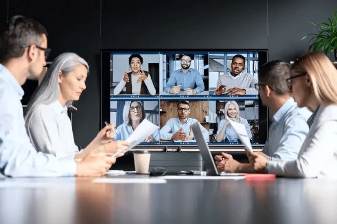Photo of four colleagues at a company around a conference table with six colleagues on a screen for a hybrid in-person and remote meeting.