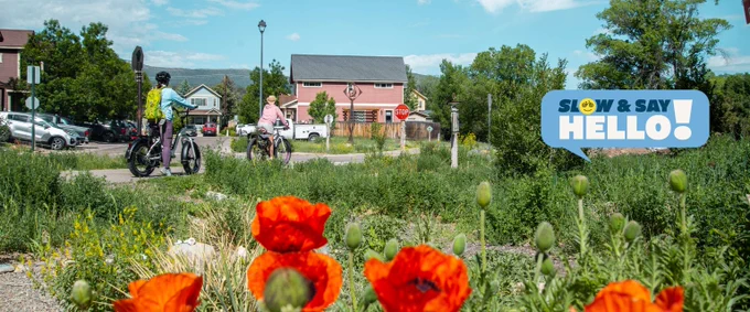 Two people riding bicycles on the Rio Grande Trail in Carbondale, Colorado with houses and greenery under a clear blue sky.
