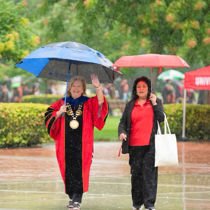 Two women walk in the rain under umbrellas. One in academic robes waves, exuding joy, while the other holds a tote bag, creating a celebratory mood.