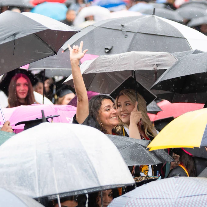 People gather closely under umbrellas in the rain, smiling and raising hands joyfully. The scene is lively, with a variety of colorful umbrellas.