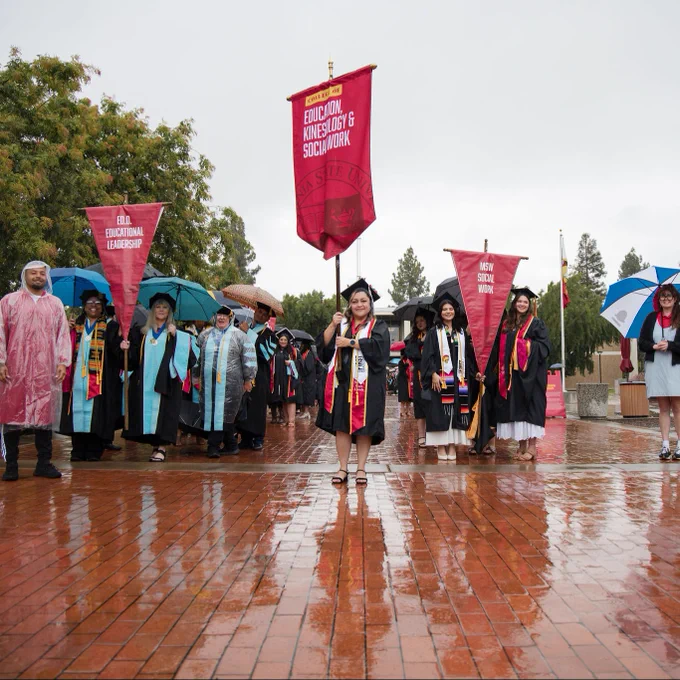 Graduates in caps and gowns stand proudly in the rain on a wet brick path, holding red banners reading "The College of Education, Kinesiology & Social Work."