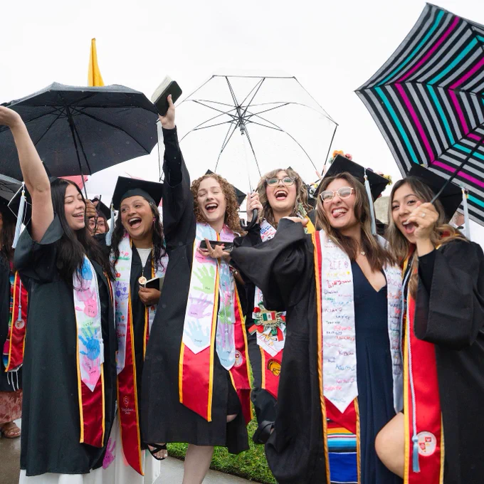 A group of graduates in caps and gowns celebrate under colorful umbrellas. They smile and cheer, conveying joy and excitement despite the rainy weather.