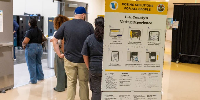 A group of individuals stands in line at a polling station. A large sign displays "L.A. County's Voting Experience" with step-by-step voting instructions. Voting booths are visible in the background.
