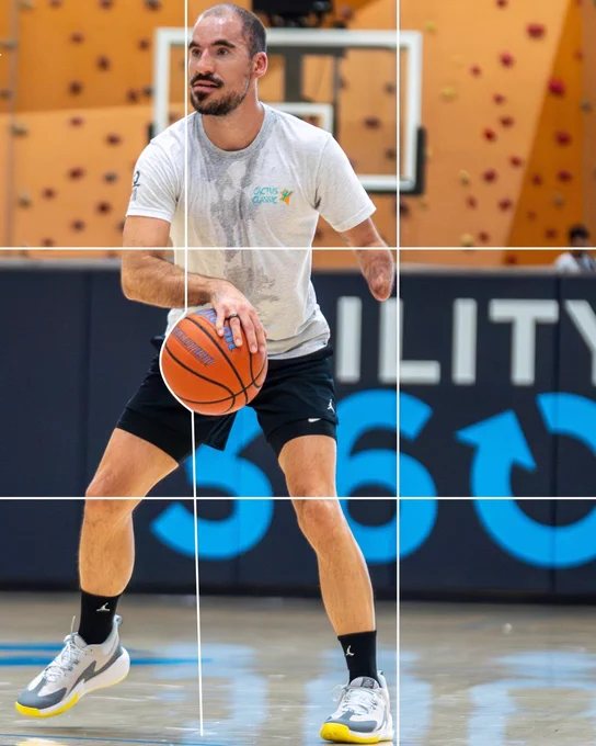 A person is playing basketball indoors, wearing a white shirt and black shorts. They are dribbling a basketball on a court. A climbing wall and basketball hoop are visible in the background.
