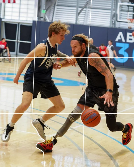 Two individuals are playing basketball on an indoor court. One person is dribbling a basketball with a prosthetic leg and wearing athletic gear. The other is attempting to block, wearing a sleeveless jersey and shorts.