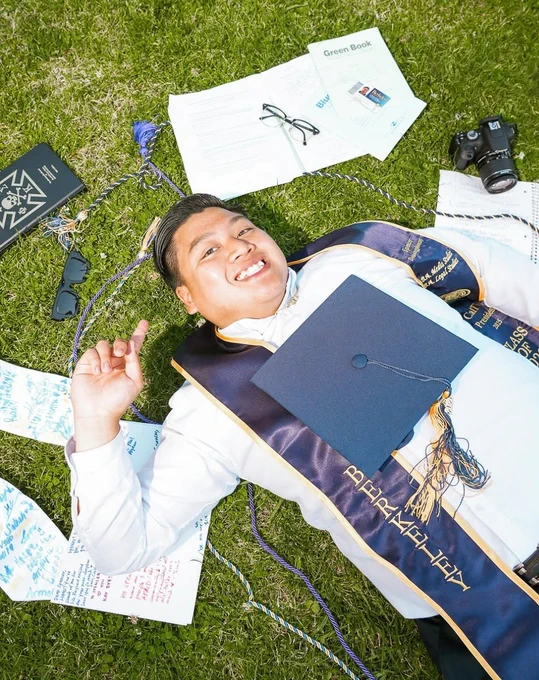 A UC Berkeley graduate poses with numerous items and their graduation cap and stole while laying in the grass of a lawn. @spencerdouangphouxay on Instagram