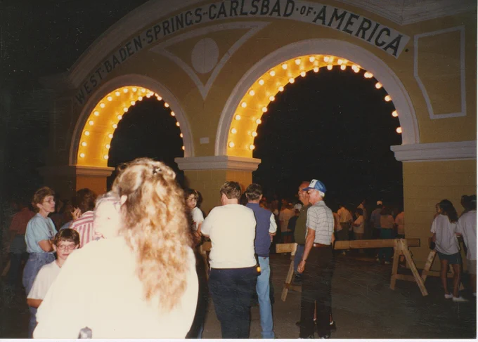 A group of people are gathered in front of an illuminated archway at night. The archway has the text "West Baden Springs-Carlsbad of America" displayed on it. The scene is lively, with many visitors milling around. The date is August 28th, 1996. The event is West Baden Springs Hotel Ice Cream Social.