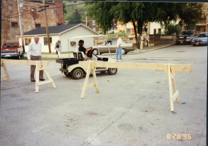 A person is driving a small utility vehicle in a parking lot, surrounded by wooden barriers. Bill Cook stands nearby, wearing a white shirt and wearing a smile of satisfaction. In the background, there are parked cars, a small building, and trees. The photo is dated August 28, 1996.