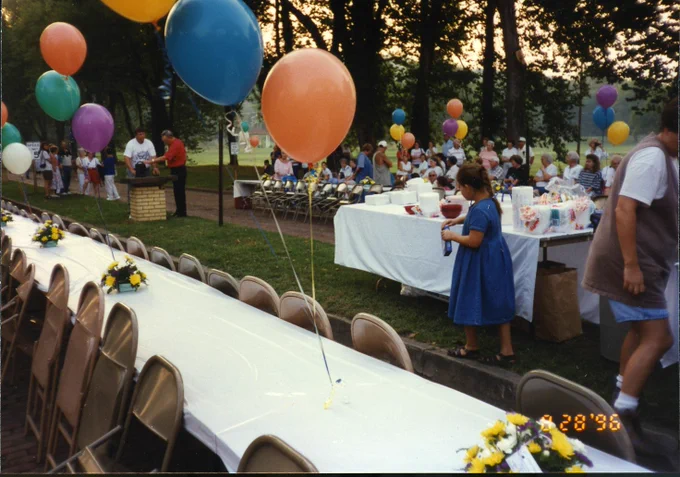 A large outdoor gathering with tables covered in white cloth and decorated with colorful balloons and floral arrangements in celebration of the relighting of West Baden Springs Hotel archway. Several people are present, some seated, others standing near the tables. In the background, trees and a park-like setting are visible. August 28, 1996 Ice Cream Social.