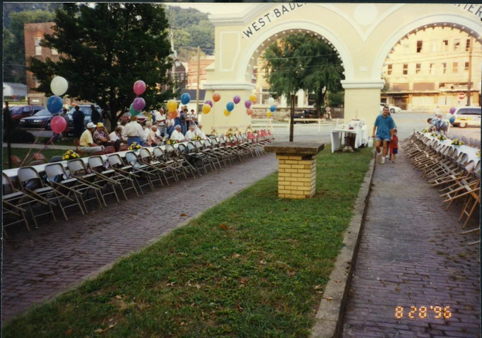 An outdoor event with rows of chairs arranged along a brick pathway under the "West Baden Springs Hotel" arch. Tables are set up with refreshments, and colorful balloons are tied to some chairs. People are gathered, and a person walks with a child. The surrounding area features grass, trees, and distant buildings.  August 28, 1996 Ice Cream Social.