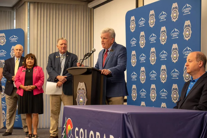 Group of officials standing and sitting during a press conference with Colorado Springs Police backdrop and a speaker at the podium.