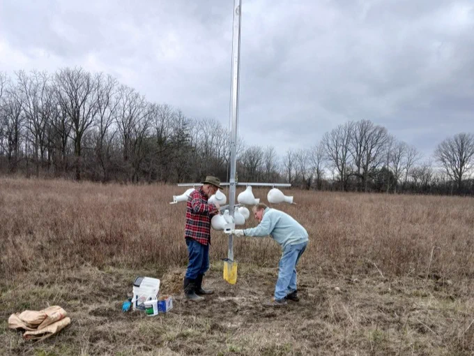 Two people work on a weather station in a grassy field. The sky is overcast, and leafless trees are visible in the background. Some tools and equipment are on the ground nearby.
