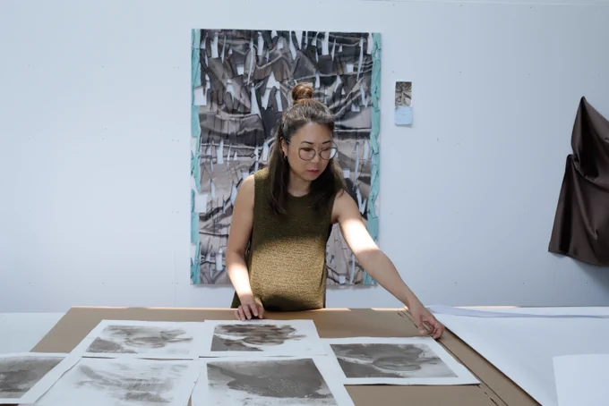 An artist wearing glasses and an olive-colored sleeveless tank top leans over a work table, examining several grayscale prints and artwork samples. Behind them, a turquoise-framed abstract artwork with black and white geometric patterns hangs on a white gallery wall.