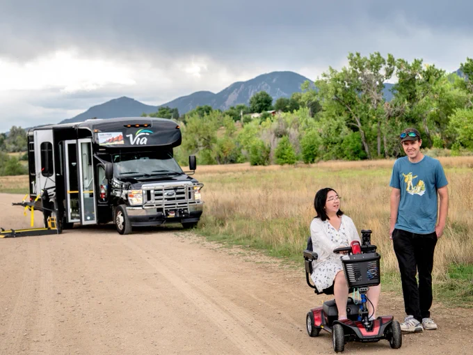 A man stands next to a woman in a motorized wheelchair along a dirt road. In the background is a parked black van with a wheelchair ramp extended out. Trees, mountains and a field frame the subjects.