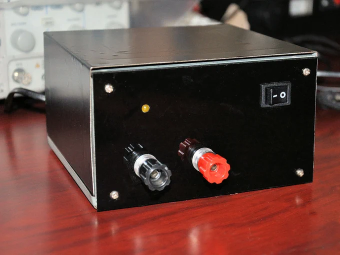 Close-up photo of a simple black bench power supply sitting on a desk, with red and black binding posts, a small power switch, and blurred lab equipment in the background.