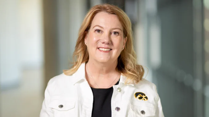 A woman smiles in a professional portrait while wearing a white Iowa Hawkeyes jacket over a black top. The softly blurred indoor background features large windows and neutral tones.