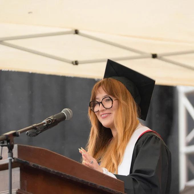 A graduate in cap and gown speaks at a podium under a tent. She is smiling, wearing glasses, and exudes confidence and joy during the ceremony.