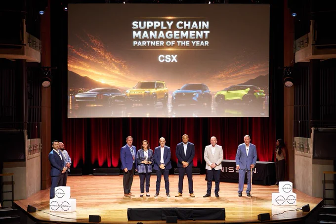 A group of individuals stands on a stage in front of a large screen displaying the text "Supply Chain Management Partner of the Year" and "CSX." The backdrop features images of cars. The stage has red curtains and Nissan logos are visible on podiums.