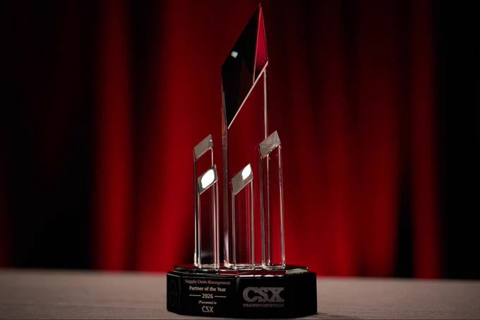 This image shows a sleek, modern glass award with a black base bearing the text "Supply Chain Management Partner of the Year 2026" and the logos for CSX and Transportation. The backdrop is a rich red curtain, adding elegance to the scene.