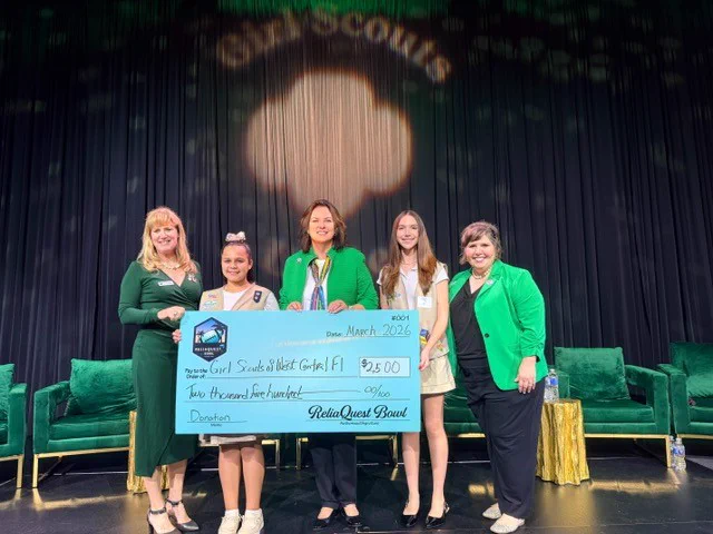 A group of people stand on stage holding a large check for the Girl Scouts of West Central Florida during a ceremony. The mood is celebratory and community-focused.