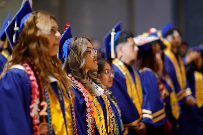 A group of graduates in blue caps and gowns stands in a row, wearing colorful stoles, during a graduation ceremony. The focus is on the person at the front, who is wearing glasses.
