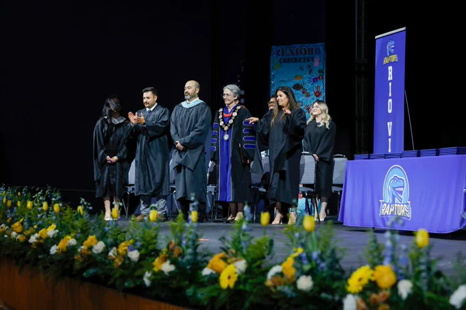A group of individuals in academic attire, including UTEP President Heather Wilson, stand on a stage decorated with floral arrangements. A table with a banner displaying "Rio Vista" and a Raptors logo is visible.
