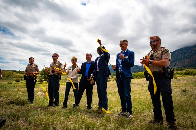 Group of seven people, including officers and officials, cutting a yellow ribbon outdoors on a grassy field under a cloudy sky.