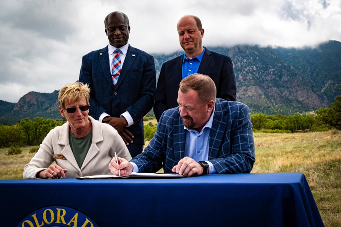 Two individuals seated at a table covered with a Colorado-branded cloth, signing documents outdoors with mountains and cloudy sky in the background, two others standing behind them.