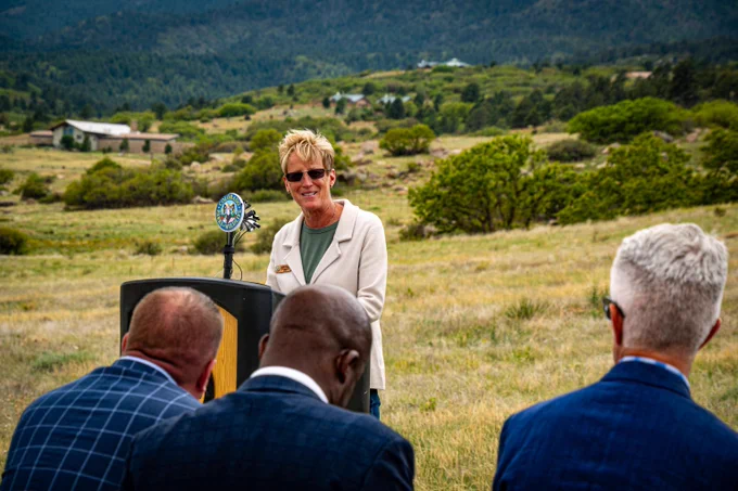 Speaker delivering a speech in a grassy field with rolling hills and trees, while three men listen attentively in the foreground.